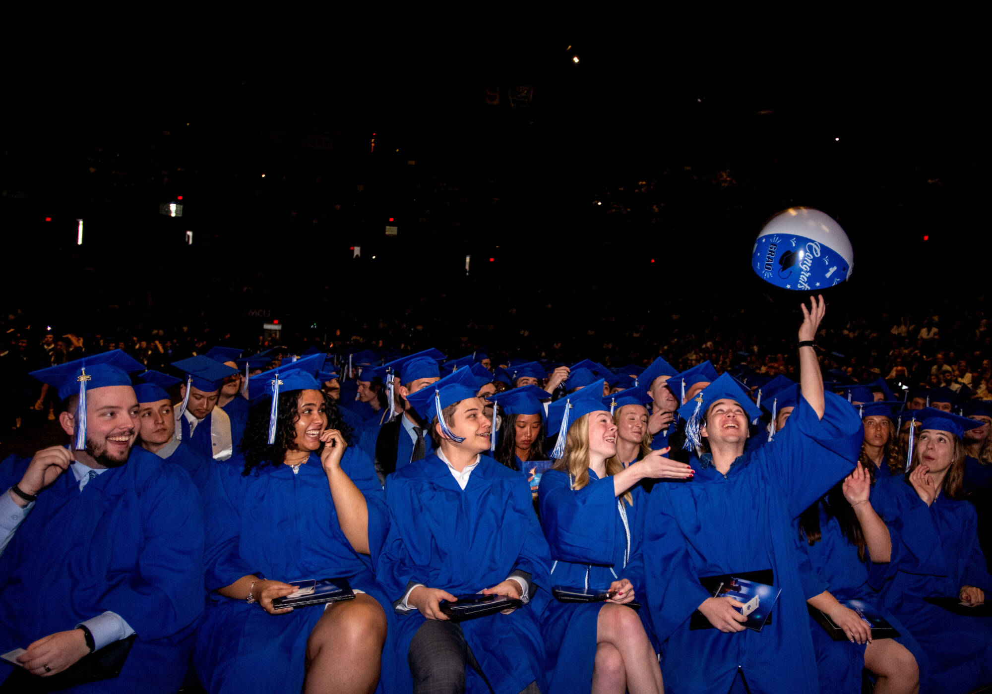 A group of students in blue graduation caps and gowns sit together during a commencement ceremony.